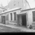 Image: A low, rectangular stone and brick building surrounded on three sides by much taller brick and stone buildings. The back door of the building features a wooden latticework awning