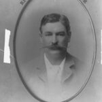 Image: A photographic head-and-shoulders portrait of a man in suit and tie sporting a large handlebar moustache. The frame surrounding the portrait identifies the man as Richard Chaffey Baker