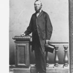 Image: A man with a chin-beard and wearing mid-Victorian era attire poses for a photograph. He stands next to a bannister and is holding a stove-pipe hat