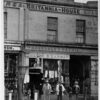 Image: three men stand outside a drapery shop in part of a two storey terrace building. A figurine of a woman holding a trident can be seen above the door to the shop.