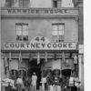 Image: A group of men and children stand outside a two storey terrace building the ground floor of which is serving as a drapery store with fabric sitting in baskets on the street, coats hanging along the storefront and hats in the window.