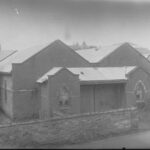 Image: An old stone and brick building with two stained glass windows and exterior stone fence