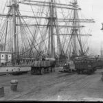 Image: Horse-drawn carts containing numerous large sacks of flour travel along a wharf next to a large, three-masted sailing ship