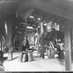 Image: The interior of a large warehouse with belt-driven machinery. Full gunny sacks are arranged near the machinery, and one sack is positioned beneath a large funnel