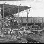 Image: A group of men use wheelbarrows to move large sacks of wheat from a shed. Within the shed are numerous tall stacks of wheat sacks