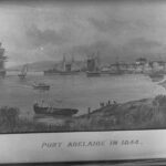 Image: A number of sailing ships are moored in a river. A scattering of buildings and a wharf are visible in the background