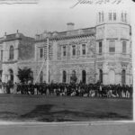 Image: A large group of men dressed in late nineteenth century attire pose for a photograph in front of a large, two-storey stone building with a corner octagonal tower. A dirt street is visible between the photographer and the group of men