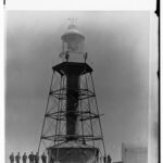 Image: A lighthouse stands at the top of a hill on a desolate island. A group of men in early Edwardian apparel pose for a photograph next to and in front of the lighthouse tower