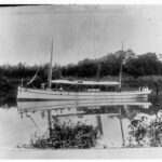 Image: Two Caucasian men stand in the bow of a late-nineteenth century steamboat as it travels down a placid river. The name ‘Victoria’ is painted on the bow of the vessel