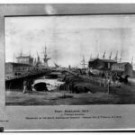 Image: A shallow creek contains two boats, one upturned. A small group of Aboriginal people sit in the foreground. On the right is a busy street with horse drawn wagons taking bales to be loaded onto ships in the background