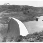 Image: A large concrete dam located among rolling hills. To the right of the dam is a reservoir of water, and to its left a dry rocky chasm