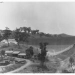 Image: A large concrete dam located among rolling hills looms behind a group of small timber buildings