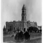 Image: A large, two-storey stone building with a tall clock tower protruding from one corner. A tree-lined park is visible in the foreground