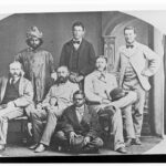 Image: A group of seven men, including an Afghan cameleer and Aboriginal tracker, pose for a photograph. The cameleer is dressed in ornate traditional costume with a turban