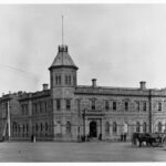 Image: A large, two-storey stone building in Victorian Italianate style. It features a large, octagonal tower with an additional storey in one corner. Two horse-drawn carts are present in the street in front of the building