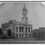 Image: A large stone building with a central tower capped by a dome. Several flags protrude from windows within the tower