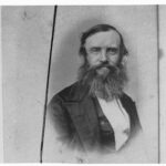 Image: A middle-aged man with a long, unkempt beard and dressed in mid-nineteenth century attire poses for a photograph