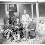 Image: black and white photograph of six men, three seated and three standing in front of a building. Kingston is seated in the front middle and wears a flower in his lapel.