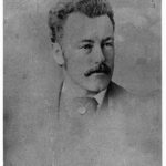 Image: A photographic head-and-shoulders portrait of a young, moustachioed man dressed in Victorian attire. He is wearing a cravat with a large pin or pendant embedded within it