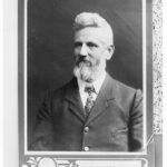 Image: A photographic portrait of a middle-aged Caucasian man with a full beard and head of hair. He is wearing an Edwardian-era suit and tie