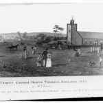 Image: a black and white copy of a watercolour painting which shows men, women and children in 1840s dress walking or riding in horse drawn carts towards a simple stone church with a single tower which is surrounded by cottages.