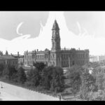 Image: A large, two-storey stone building with a tall clock tower protruding from one corner. A tree-lined park is visible in the foreground