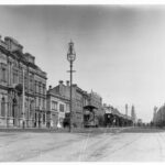 Image: black and white photograph of city street, with tram, horse drawn vehicles and pedestrians