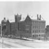 Image: A large, five storey, brick and stone building with a central tower, bay windows, crenellated parapet and pitched iron roof. The dome of the exhibition building can be seen behind.