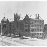 Image: A large stately building made of brick fronted by a large street