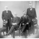 Image: A group of middle-aged men in Edwardian attire pose for a photograph. The two men in the centre sit on chairs, while the other two stand on either side of the group