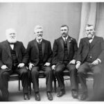 Image: A group of middle-aged Caucasian men in Edwardian-era suits pose for a photograph seated on leather seats