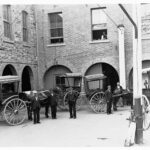 Image: Five men in uniforms stand in front of three horse-drawn carts in a building courtyard. Painted on the side of the carts are the words ‘J. Reid & Co. Mail Contractors, 42 Waymouth St.’