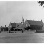 Image: A group of children stand on the street outside a walled symmetrical stone building with a verandah with stone arches, a central spire, and two gable roofed wings with rose windows on the facade and double arched windows along the sides.