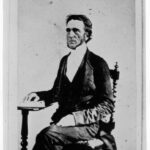 Image: Photographic portrait of a middle-aged Caucasian man in the clothing of a Christian minister. He is sitting in an high-backed chair and has his right hand resting on a book--possibly a bible