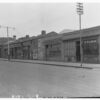 Image: a terrace of mostly single storey commercial buildings (one double storey building can be seen) including a 2nd hand store which has coats hanging outside, a boot maker and a furniture manufacturer which has boarded up windows.