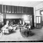 Image: A group of small children play in a large open room while four female teachers in Edwardian attire look on