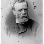 Image: A photographic head-and-shoulders portrait of a bearded, middle-aged man wearing a suit with cravat and jewelled cravat pin