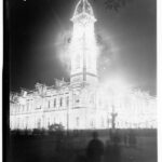 Image: A large, two-storey building with a tall clock tower in one corner is brilliantly lit at night. A small number of people on the street below look at the building