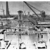 Image: The supporting framework of a bridge under construction extends across a river. Sailing ships, a wharf, and a large brick smokestack are visible in the image background