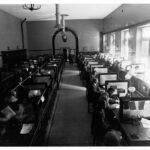 Image: A group of men sit at two rows of tables in a large open room. Each man has telegraph equipment on the table in front of him