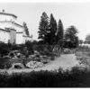 Image: An ornate conservatory building sits within a densely planted garden in full bloom with a winding gravel path in the foreground.