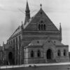 Image: A stone hall with buttresses and arched windows. Two entrances can be seen, one at the front and one at the side which both lead into vestibules.