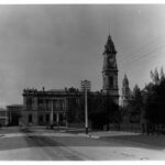 Image: A large, two-storey stone building with a tall clock tower protruding from one corner. A tree-lined park with wrought-iron fence is visible in the right foreground