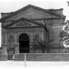 Image: a windowless classical revival style stone building behind a stone and wrought iron wall/fence