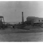 Image: A cylindrical brick chimney is surrounded by a number of buildings and industrial machinery