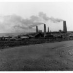 Image: Several large brick chimneys are flanked by a number of buildings. A vague coastline is visible in the background