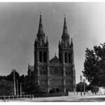 Image: A large, Gothic stone cathedral with two tall spires and a central window with floral motif