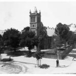 Image: A large, stone church surrounded by trees stands on the corner formed by two streets. A man in a brimmed hat stands in front of the churchyard