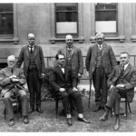 Image: Six men in suits pose for a photograph in front of a large building. Three men are seated in the front row, while the remainder stand in another row behind