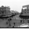 Image: A view from a busy intersection with many people in 1920s attire walking or cycling, and a mix of horse drawn and motor vehicles. The view is centred on a street lined with 2 and 3 storey commercial buildings, most with verandahs and/or balconies.